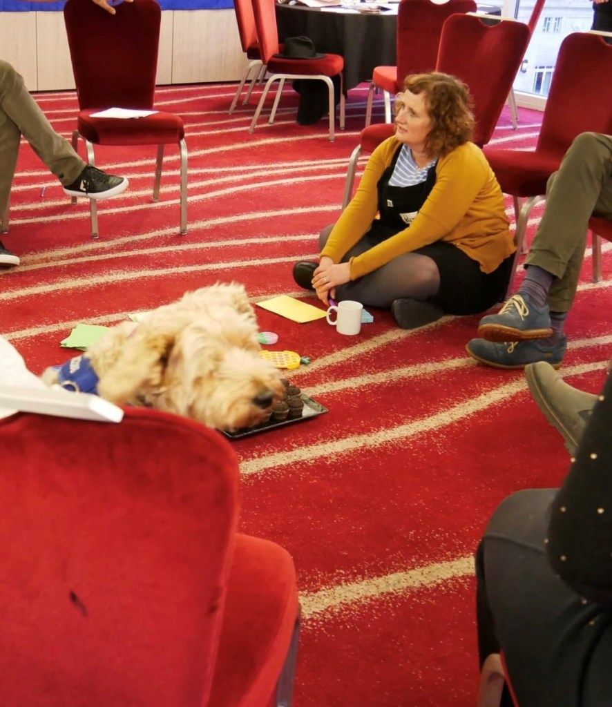Photo of Ruth Beattie and a dog. Sitting on floor listening.